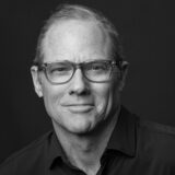 Black and white photo of Will Warne wearing a black button down shirt and glasses against a dark backdrop.