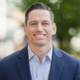Chris Rye standing outside, smiling at the camera in a blue blazer and blue button down shirt.