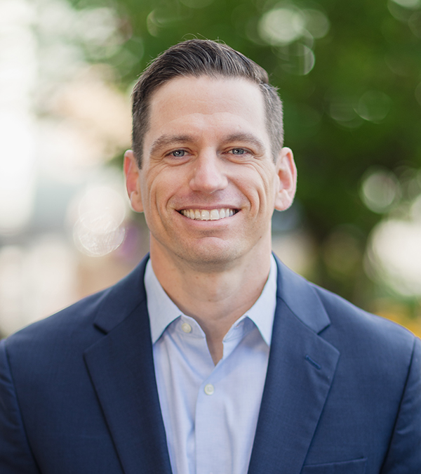 Chris Rye standing outside, smiling at the camera in a blue blazer and blue button down shirt.